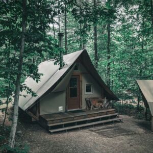 A small, A-frame cabin with canvas walls and a wooden porch nestled in a dense green forest.
