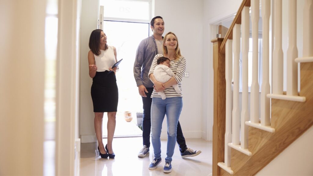 Three adults and a baby standing in a home entryway, with one adult holding a folder and gesturing while the others look upward