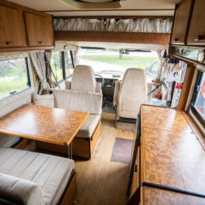 The interior of a recreational vehicle featuring a wooden dining table, beige upholstered bench seating, and overhead cabinets.