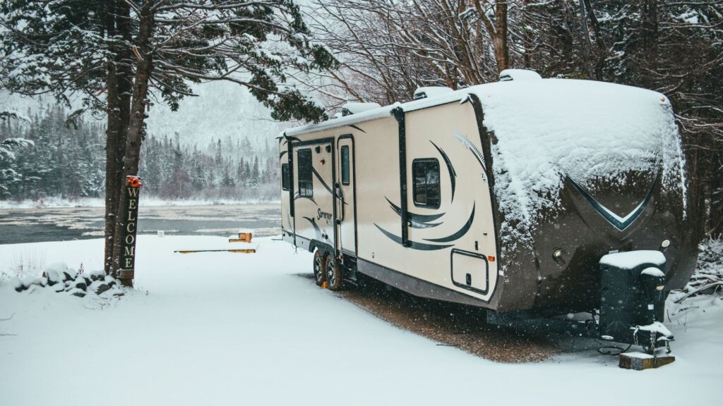 A beige travel trailer covered in a layer of snow parked in a winter setting.