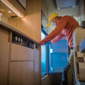 A person wearing a yellow cap bending down to look inside an open refrigerator unit.